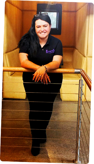 Smiling woman wearing a black polo shirt, leaning on a railing in a softly lit interior space with padded walls.