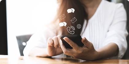 A woman holding and tapping a smartphone at a table, with floating icons representing messages, email, phone calls, and social media above the device.